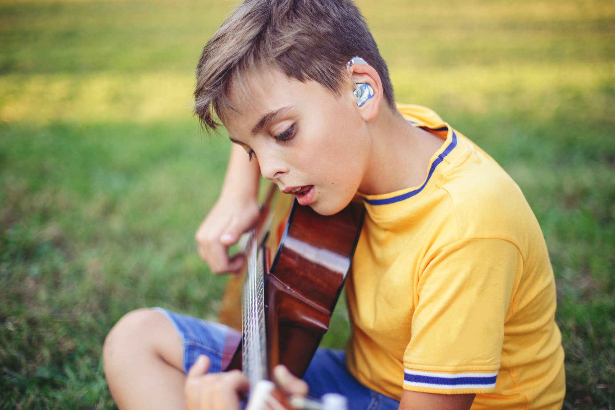 Hard of hearing preteen Caucasian boy with hearing aids playing guitar and singing outdoor